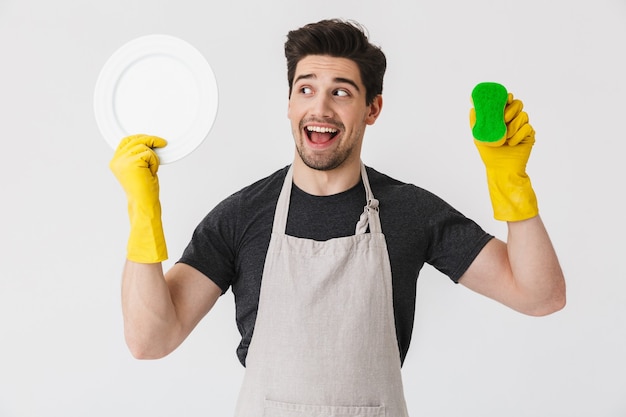 Foto de um jovem animado usando luvas de borracha amarelas para proteção das mãos, lavando pratos enquanto limpa a casa isolada sobre o branco