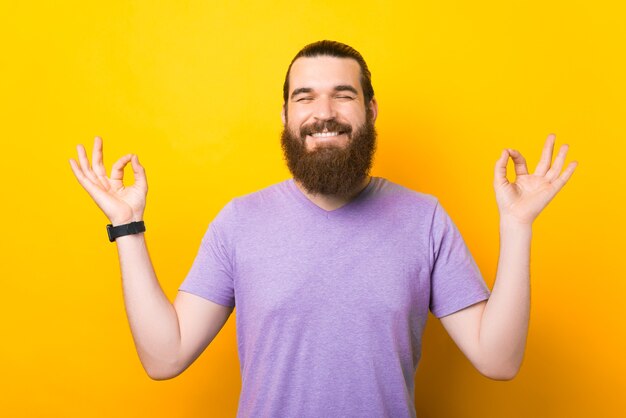 Foto de um jovem barbudo alegre e feliz em pé sobre um fundo amarelo e fazendo um gesto zen, meditando