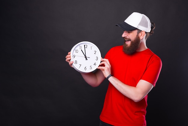 Foto foto de um jovem barbudo com uma camiseta vermelha e boné branco segurando um relógio às onze horas