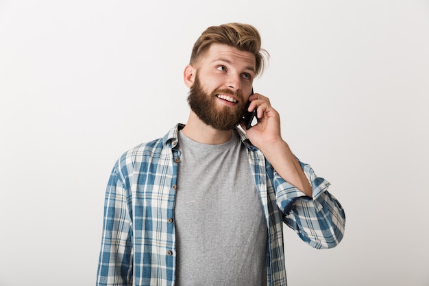 Foto de um jovem bonito barbudo em pé, isolado sobre o fundo da parede branca, falando por telefone celular.