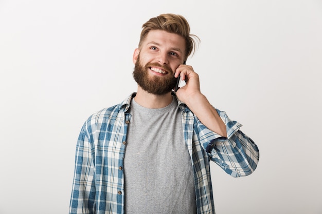 Foto de um jovem bonito barbudo em pé, isolado sobre o fundo da parede branca, falando por telefone celular.