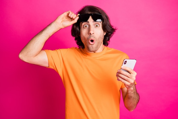 Foto de um jovem braço descolando óculos segurar telefone usar camiseta laranja óculos de sol isolado fundo de cor rosa