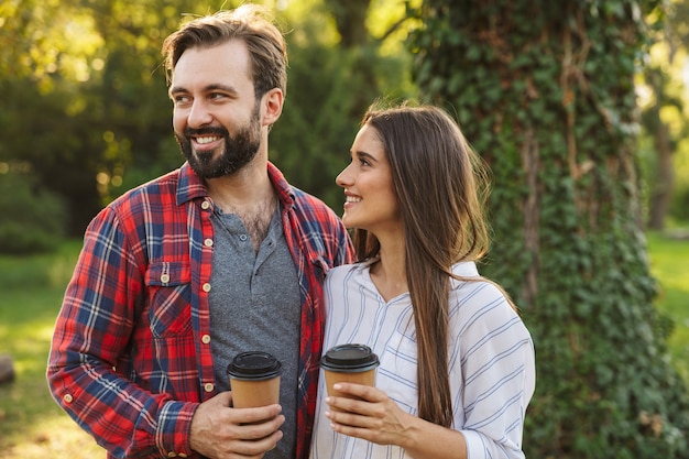 Foto de um jovem casal apaixonado, alegre, caminhando ao ar livre em uma floresta verde do parque natural, bebendo café.