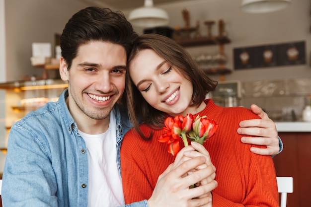 Foto de um jovem casal apaixonado, homem e mulher, segurando lindas flores juntos, enquanto está sentado à mesa em um café aconchegante