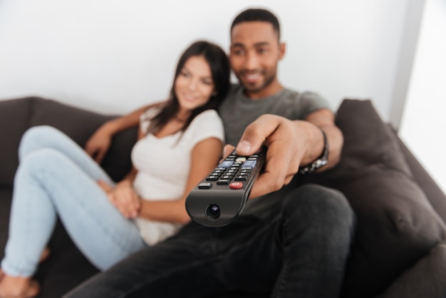 Foto de um jovem casal feliz abraçando e assistindo tv no sofá em casa. foco disponível com controle remoto.