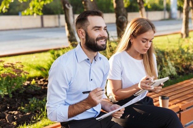 Foto de um jovem concentrado otimista positivo homem e mulher empresários do lado de fora no parque natural escrevendo notas em cadernos.