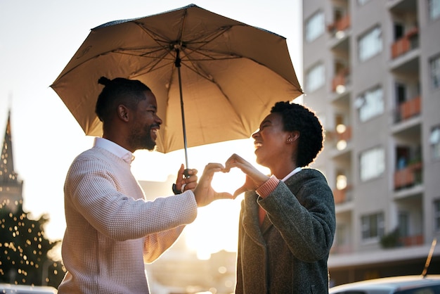 Foto de um jovem e uma mulher de pé debaixo de um guarda-chuva e fazendo um gesto em forma de coração enquanto caminhava na chuva contra um fundo urbano