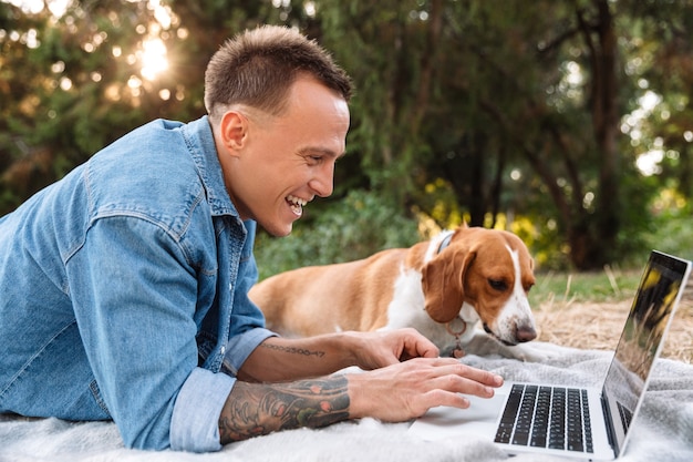 Foto de um jovem feliz deitado em um cobertor no parque com seu cachorro canino e o computador laptop