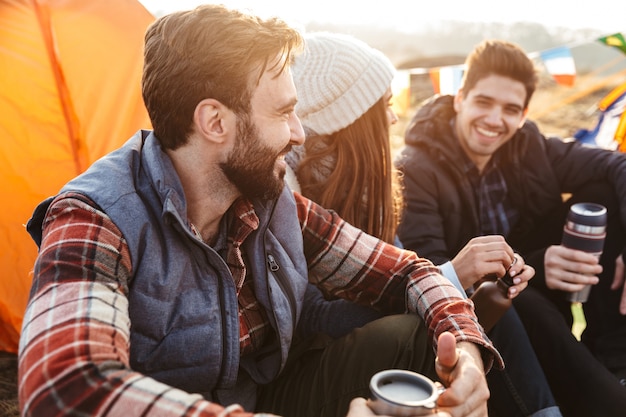 Foto de um jovem grupo de amigos do lado de fora em um acampamento de férias alternativo gratuito nas montanhas, bebendo chá quente, conversando entre si.