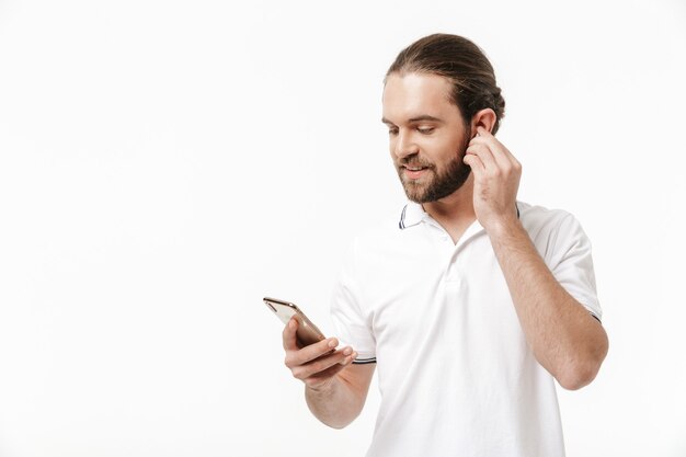 Foto foto de um jovem homem barbudo bonito alegre posando isolado sobre uma parede branca, usando telefone celular ouvindo música com fones de ouvido.