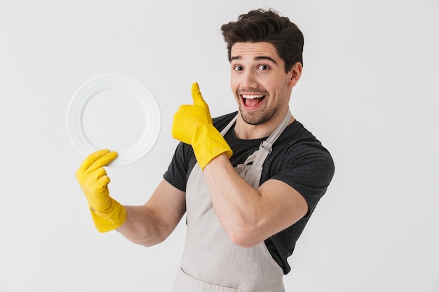 Foto de um jovem moreno usando luvas de borracha amarelas para proteção das mãos, lavando pratos enquanto limpa a casa isolada sobre o branco