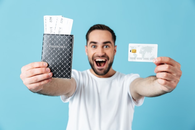 Foto de um jovem moreno vestindo uma camiseta básica, sorrindo, segurando as passagens com passaporte e cartão de crédito isolado sobre o azul