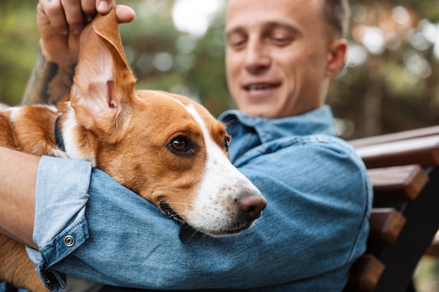 Foto de um jovem satisfeito em roupas jeans, sorrindo e sentado no banco do parque com seu cachorro