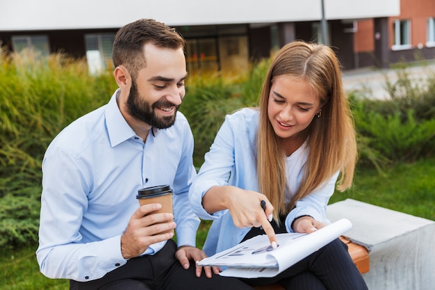 Foto de um jovem sorridente positivo homem e mulher empresários lá fora na rua segurando a prancheta com planos de negócios de documentos falando uns com os outros, bebendo café.