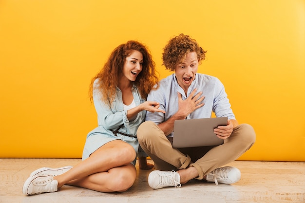 Foto de um lindo casal maravilhado, homem e mulher, sentados no chão e usando um laptop prateado, isolado sobre um fundo amarelo