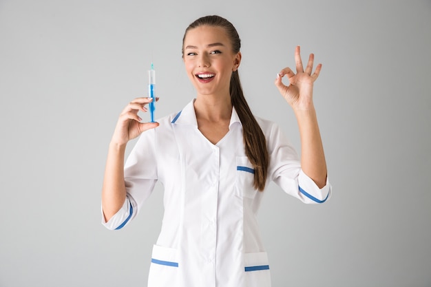 Foto de um médico cosmetologista linda jovem isolada sobre uma parede cinza segurando a seringa de injeção mostrando o gesto de tudo bem.