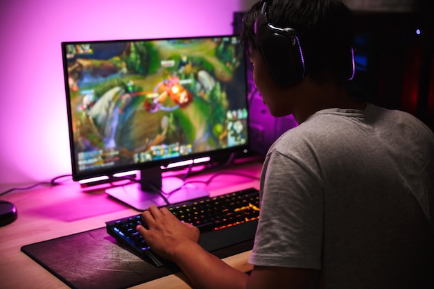 Foto de um menino adolescente jogando videogame no computador em um quarto escuro, usando fones de ouvido e um teclado colorido com luz de fundo