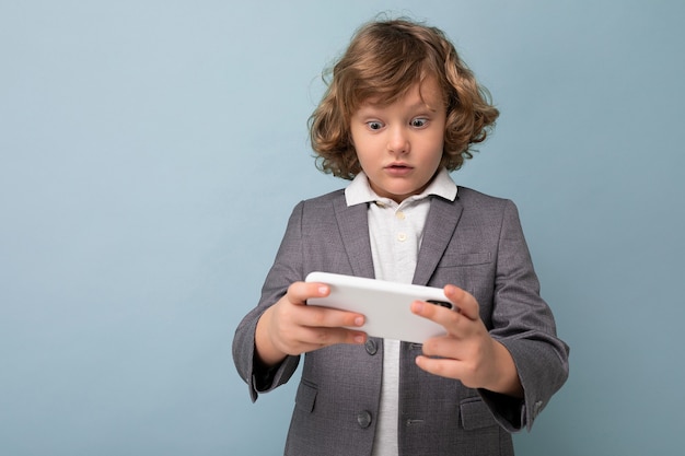 Foto de um menino bonito e emocional com cabelo encaracolado, vestindo um terno cinza, segurando e usando o telefone