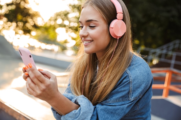 Foto de um sorridente alegre positivo jovem adolescente do lado de fora no parque, ouvindo música com fones de ouvido, segurando o telefone móvel.
