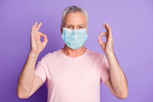 Foto de um velho aposentado levantando duas mãos mostra ok, usando máscara facial azul, fundo de cor violeta