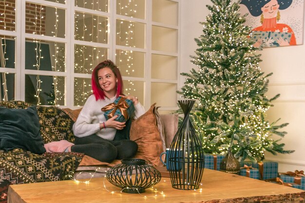 Foto de uma adorável jovem sonhadora vestida de suéter segurando um presente de Natal no interior do ano novo