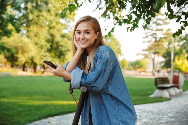 Foto de uma alegre muito bonita jovem adolescente do lado de fora no parque verde natural com scooter usando telefone celular.
