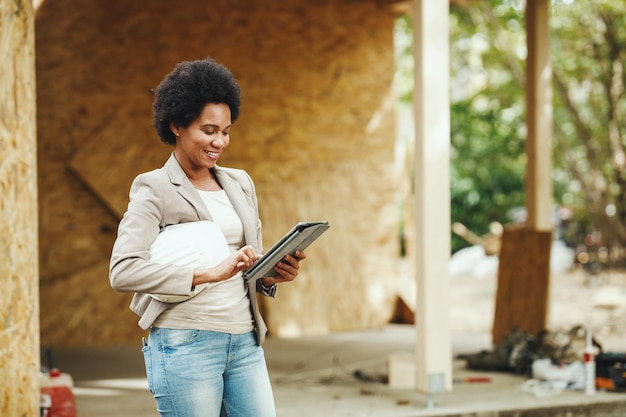 Foto de uma arquiteta africana usando um tablet digital e verificando o canteiro de obras de uma nova casa de madeira.