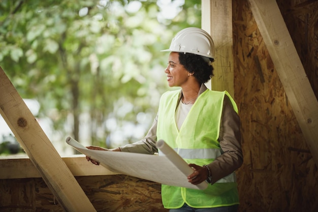 Foto de uma arquiteta africana verificando planos no canteiro de obras de uma nova casa de madeira. Ela está vestindo roupas de trabalho de proteção e capacete branco.