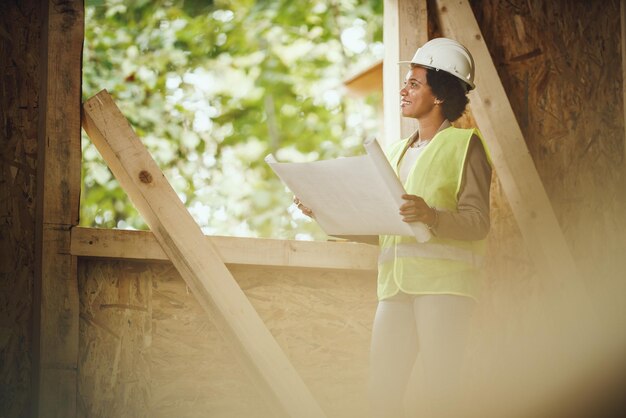 Foto de uma arquiteta africana verificando planos no canteiro de obras de uma nova casa de madeira. Ela está vestindo roupas de trabalho de proteção e capacete branco.