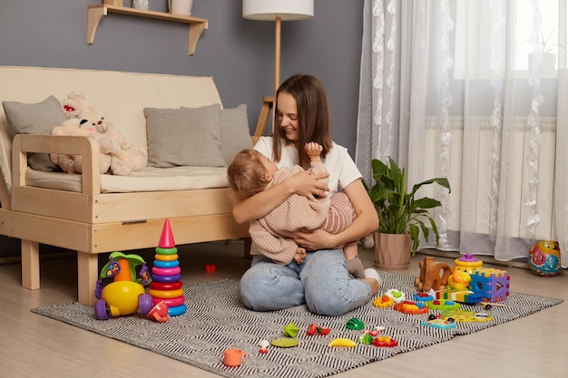 Foto de uma atraente mulher caucasiana brincando e se divertindo no chão com sua filha segurando o bebê nas mãos cantando uma canção de ninar e sorrindo expressando uma expressão positiva