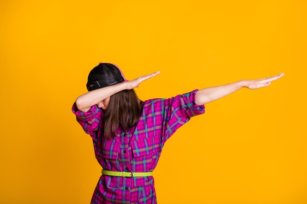 Foto de uma bela garota morena usando um boné de camisa rosa dançando e mostrando um sinal de hype dab isolado em um fundo de cor amarela