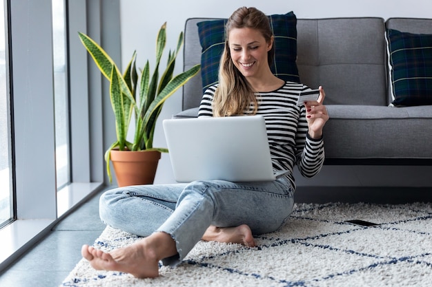 Foto de uma bela jovem compras online com cartão de crédito e laptop enquanto está sentado no chão em casa.