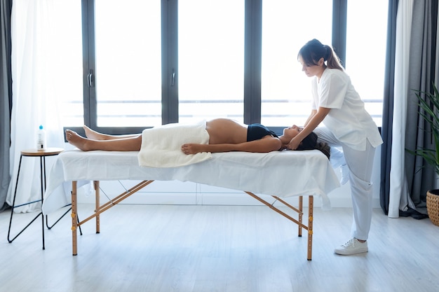 Foto de uma bela jovem fisioterapeuta fazendo tratamento osteopático ou quiroprático no pescoço de uma mulher grávida em uma maca em casa.