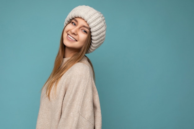 Foto de uma bela jovem loira morena sexy feliz positiva isolada sobre uma parede de fundo azul, vestindo um suéter bege quente e um chapéu de malha bege de inverno, olhando para a câmera