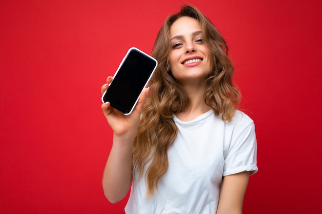 Foto de uma bela jovem loira sorridente bonita vestindo uma camiseta branca em pé isolada em um fundo vermelho com espaço de cópia segurando o telefone, mostrando o smartphone na mão com tela vazia