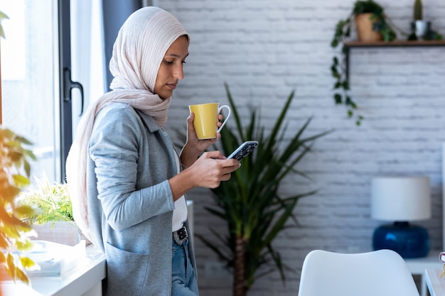 Foto de uma bela jovem mulher de negócios muçulmana usando hijab, enviando mensagens com o celular em pé perto da janela no escritório.