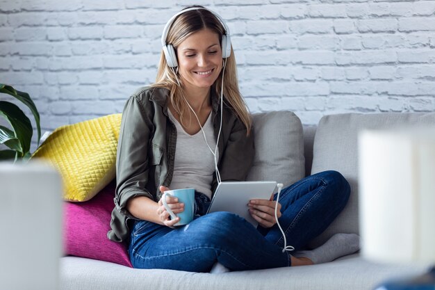 Foto de uma bela jovem ouvindo música com seu tablet digital enquanto bebia uma xícara de café no sofá em casa.