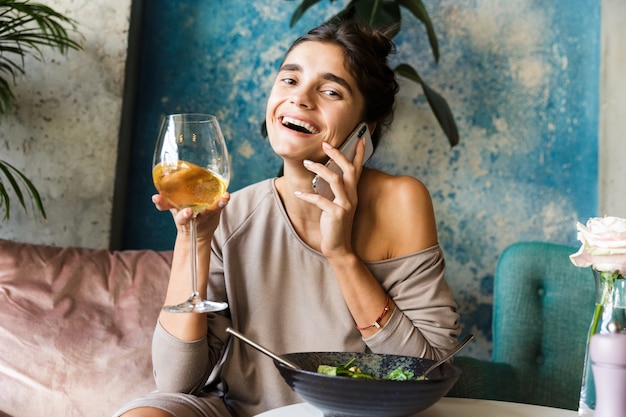 Foto de uma bela jovem sentada num café dentro de casa, bebendo vinho branco, comendo salada, falando pelo telefone celular.