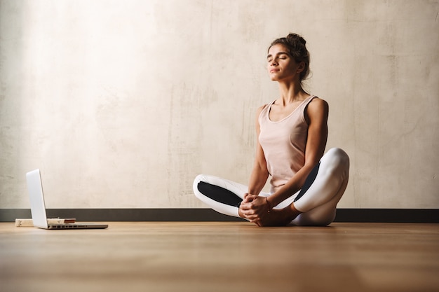 Foto foto de uma bela mulher concentrada em roupas esportivas fazendo exercícios com os olhos fechados e usando o laptop enquanto está sentada no chão em casa