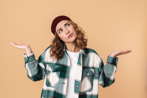 Foto de uma bela mulher pensativa com chapéu de malha, olhando para cima e segurando copyspace isolado em bege