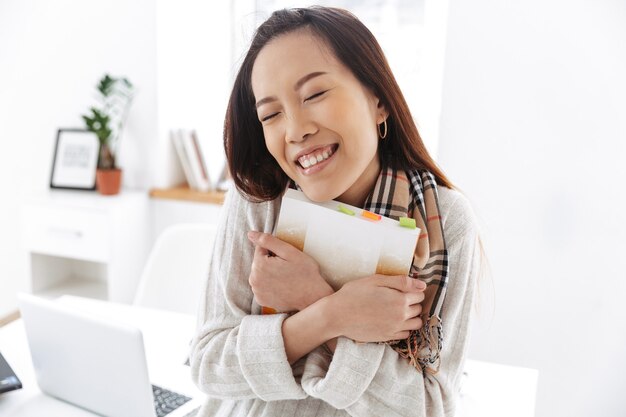 Foto de uma bela trabalhadora asiática vestindo um suéter branco segurando o diário e sorrindo enquanto está sentada na mesa no escritório