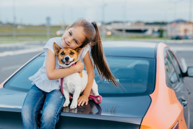 Foto de uma criança alegre e pequena tem rabo de cavalo abraça com amor seu animal de estimação favorito, posar no porta-malas do automóvel, brincar juntos, caminhar ao ar livre, desfrutar da união. Conceito de crianças e animais