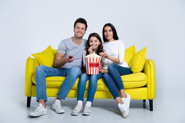 Foto de uma criança bonita e seus pais, que estão comendo pipoca juntos, enquanto assistem programas de TV no sofá.