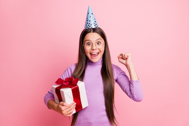 Foto de uma criança encantada segurando uma caixa de presente com os punhos levantados e usando um cone roxo do dia de festa isolado em um fundo de cor pastel