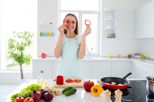 Foto de uma dona de casa encantadora aproveitando a manhã de fim de semana cozinhando um jantar saboroso segurando uma fatia de pimenta vermelha perto de um olho infantil humor despreocupado carrinho cozinha com luz branca dentro de casa