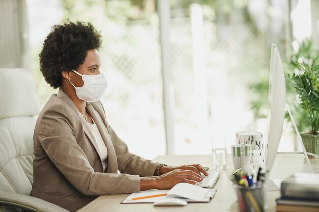 Foto de uma empresária africana com máscara protetora N95 sentada sozinha em seu escritório e trabalhando no computador durante a pandemia de COVID-19.