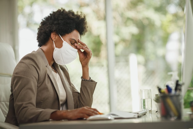 Foto de uma empresária africana estressada com máscara protetora N95 sentada sozinha em seu escritório e trabalhando no computador durante a pandemia de COVID-19.