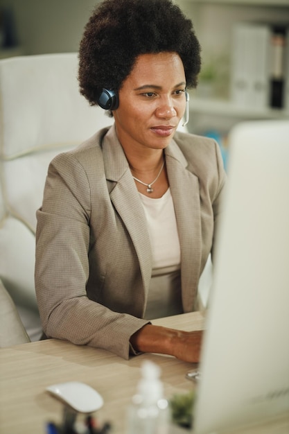 Foto de uma empresária africana usando um fone de ouvido enquanto trabalhava no computador em seu escritório durante a pandemia do vírus corona.