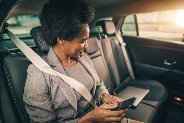Foto de uma empresária negra bem-sucedida usando smartphone enquanto está sentada no banco de trás de um carro durante seu trajeto matinal.