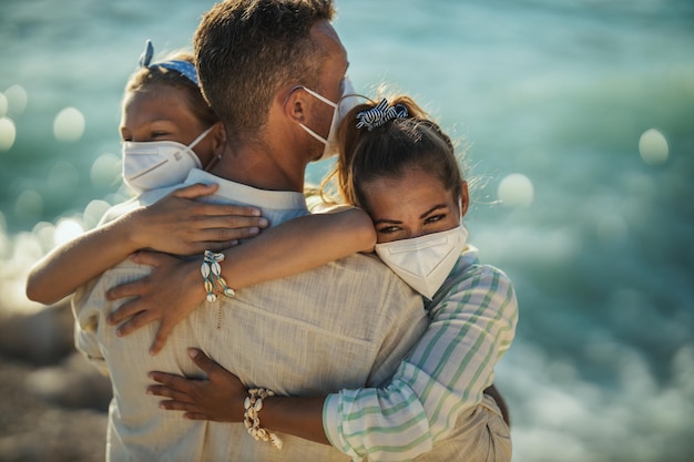 Foto de uma família feliz e abraçada com máscara protetora N95 passando tempo à beira-mar na pandemia de corona. Eles estão tendo auto-isolamento na natureza.